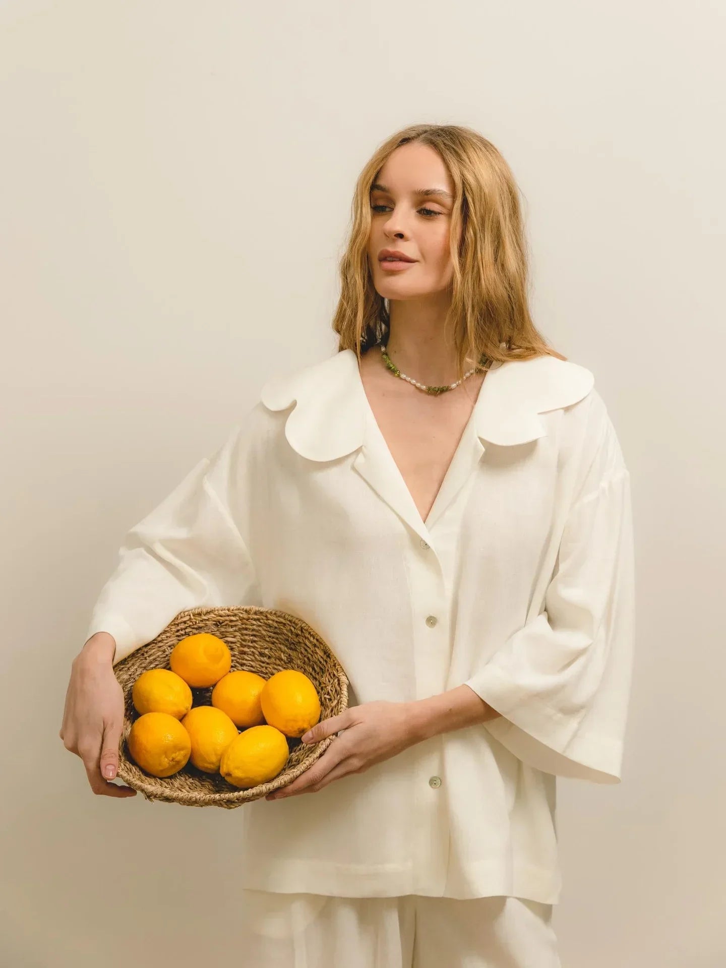 A woman with light brown hair wearing an oversized white blouse, holding a wicker basket filled with lemons, standing against a neutral background.