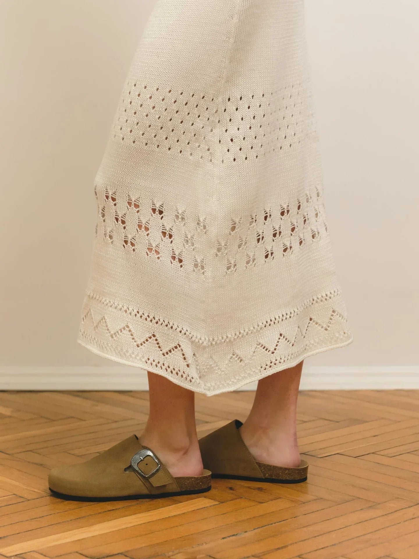 A close-up of a person wearing a cream-colored knit skirt with intricate patterns, paired with brown suede slip-on sandals, standing on a wooden floor
