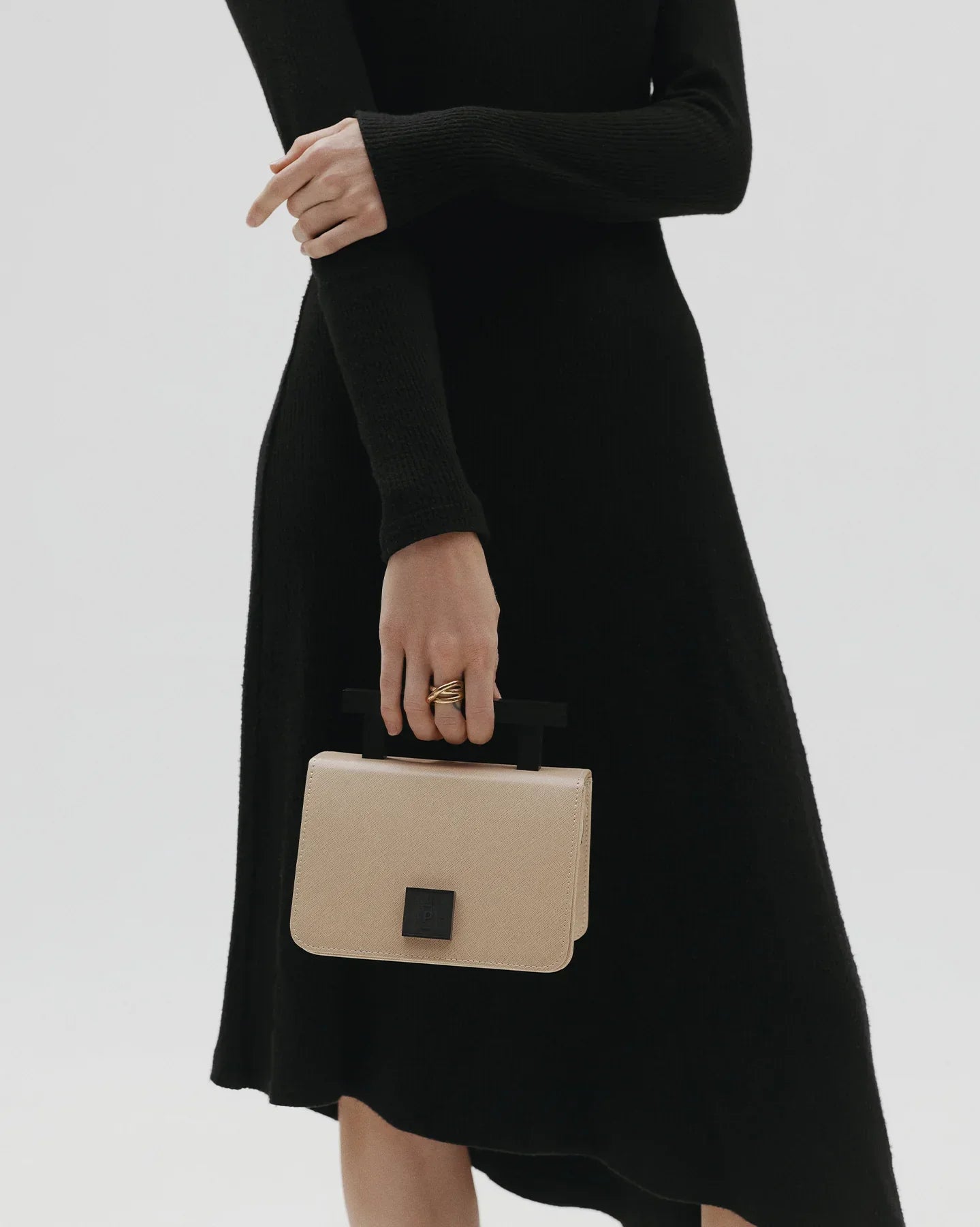 A person wearing a black dress, holding a beige handbag with a black handle, photographed from the waist down against a neutral background.