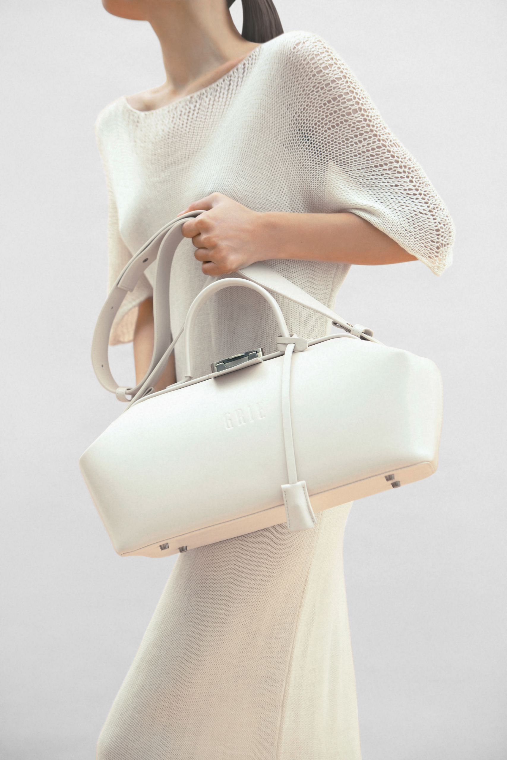 A person wearing a light beige knit dress, holding a large white handbag with a sleek design, photographed from the torso down against a neutral background.