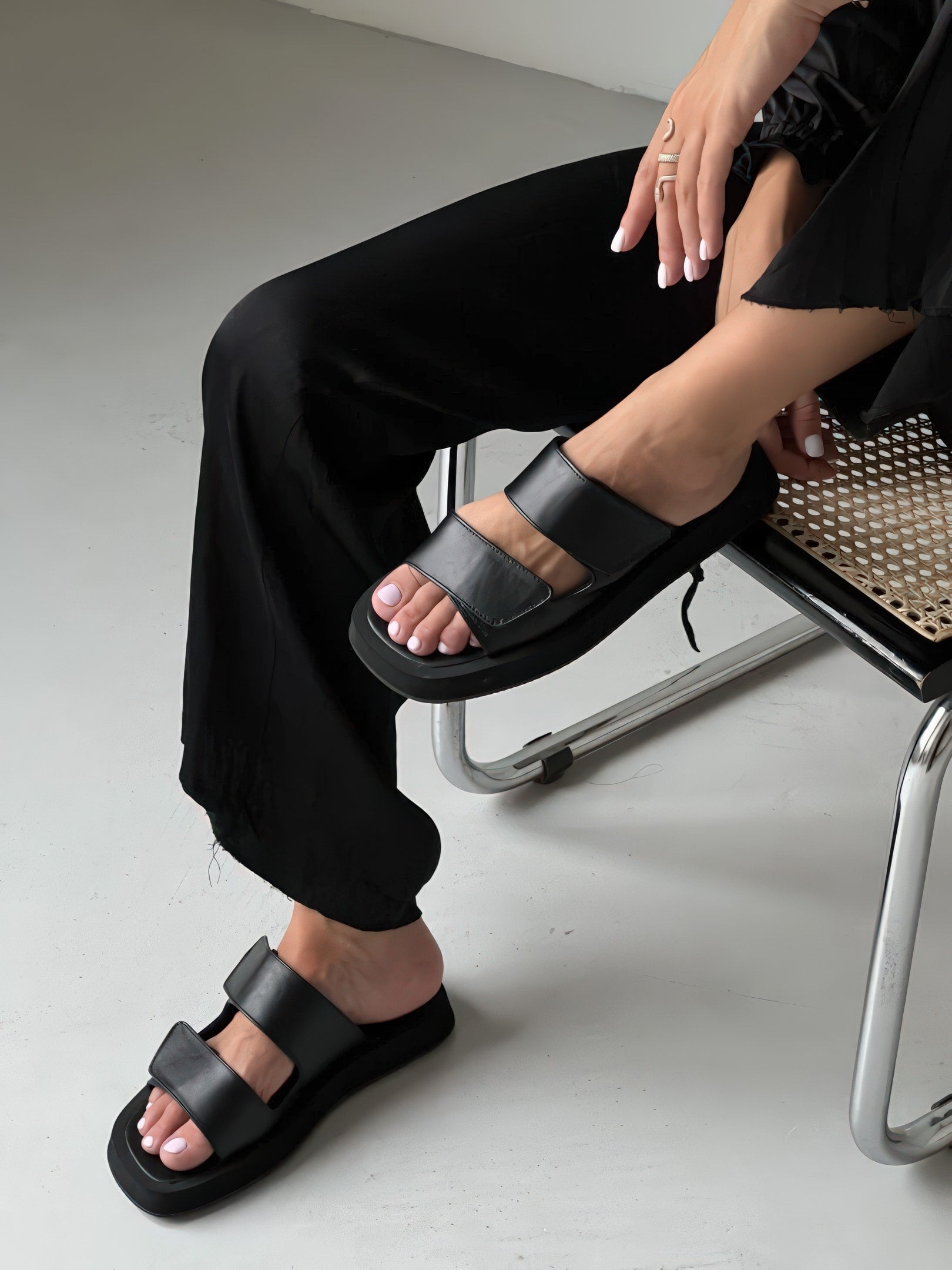 A person sitting on a chair wearing black pants and black double-strap sandals, with one leg crossed over the other, showing manicured nails.