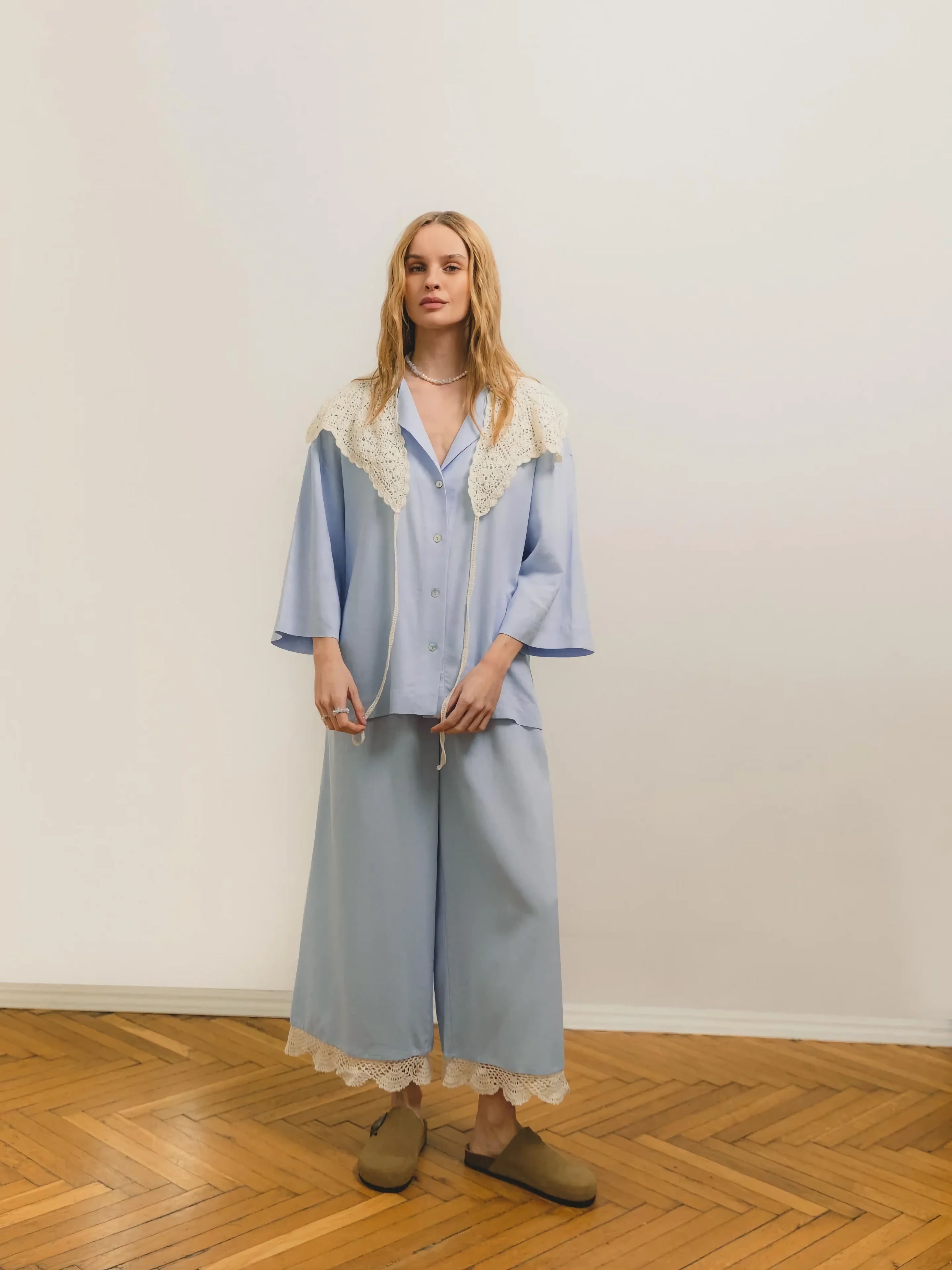 A woman with blonde hair wearing a light blue outfit featuring a button-up top with a lace collar and matching wide-leg pants with lace trim, paired with brown slip-on shoes, standing on a wooden floor against a neutral background