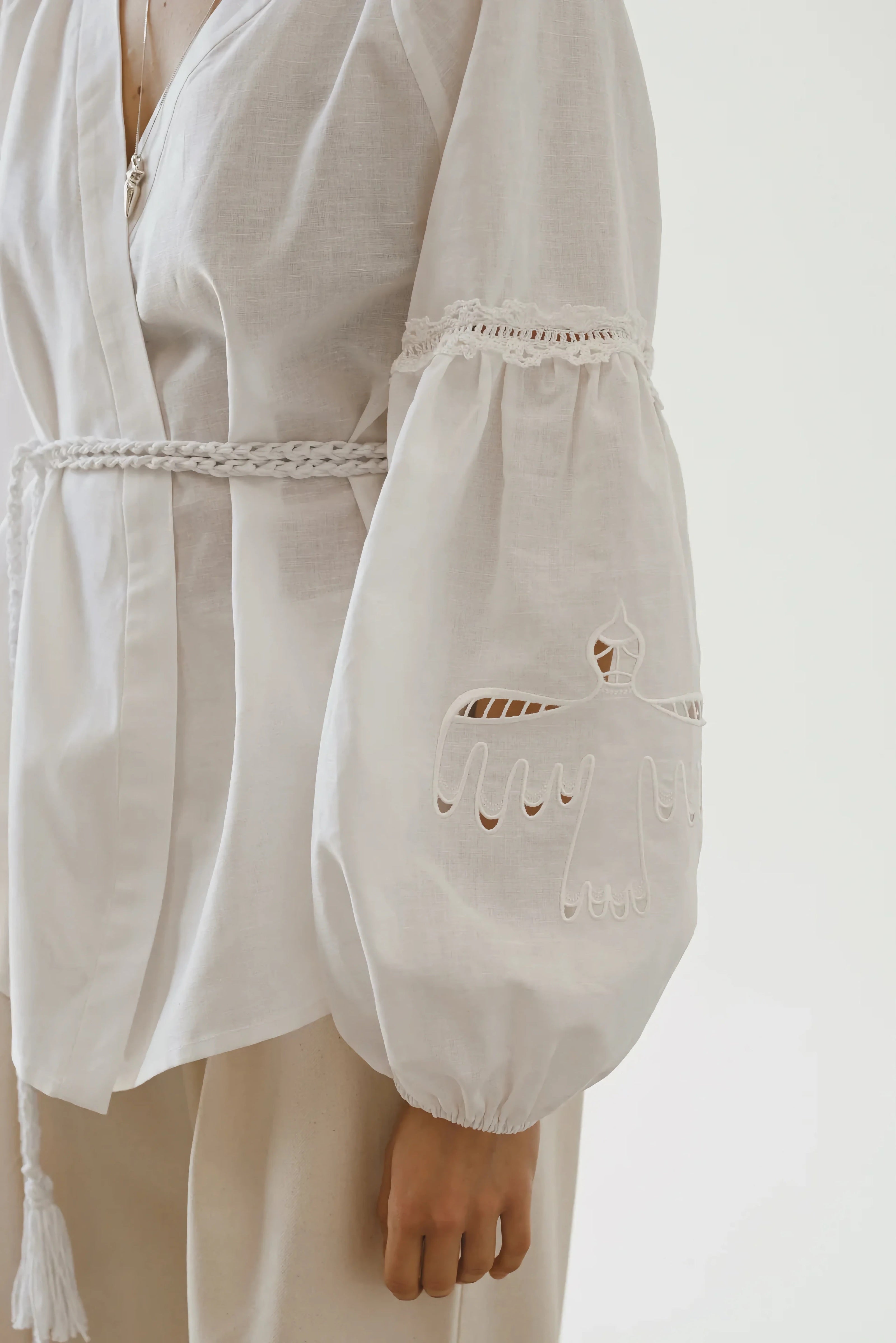 A close-up of a person wearing a white blouse with voluminous sleeves featuring intricate cut-out embroidery, paired with beige trousers, against a light background.
