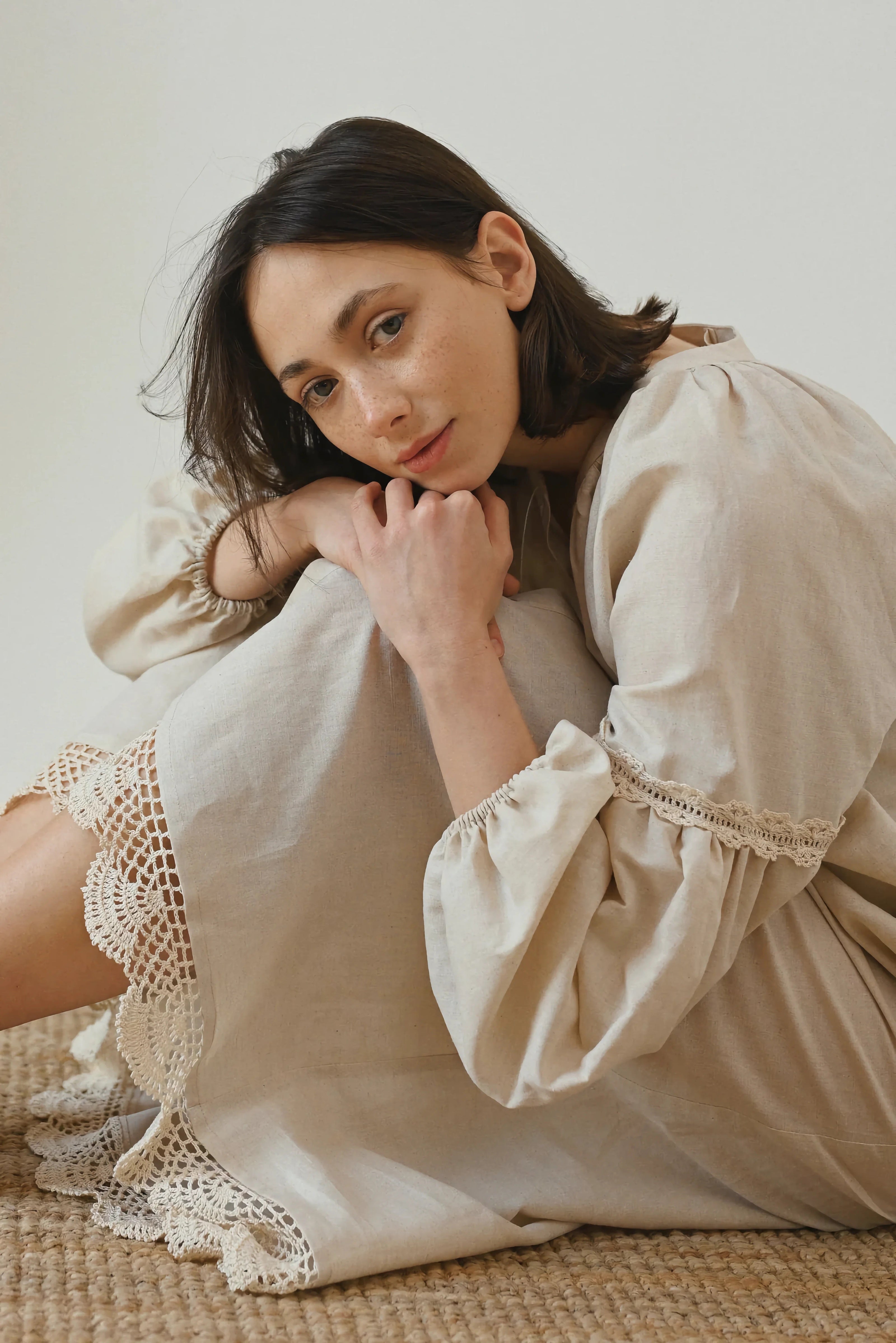A woman sitting on a woven rug, wearing a beige dress with crochet lace trim and voluminous sleeves, resting her arms on her knees and looking thoughtfully at the camera.
