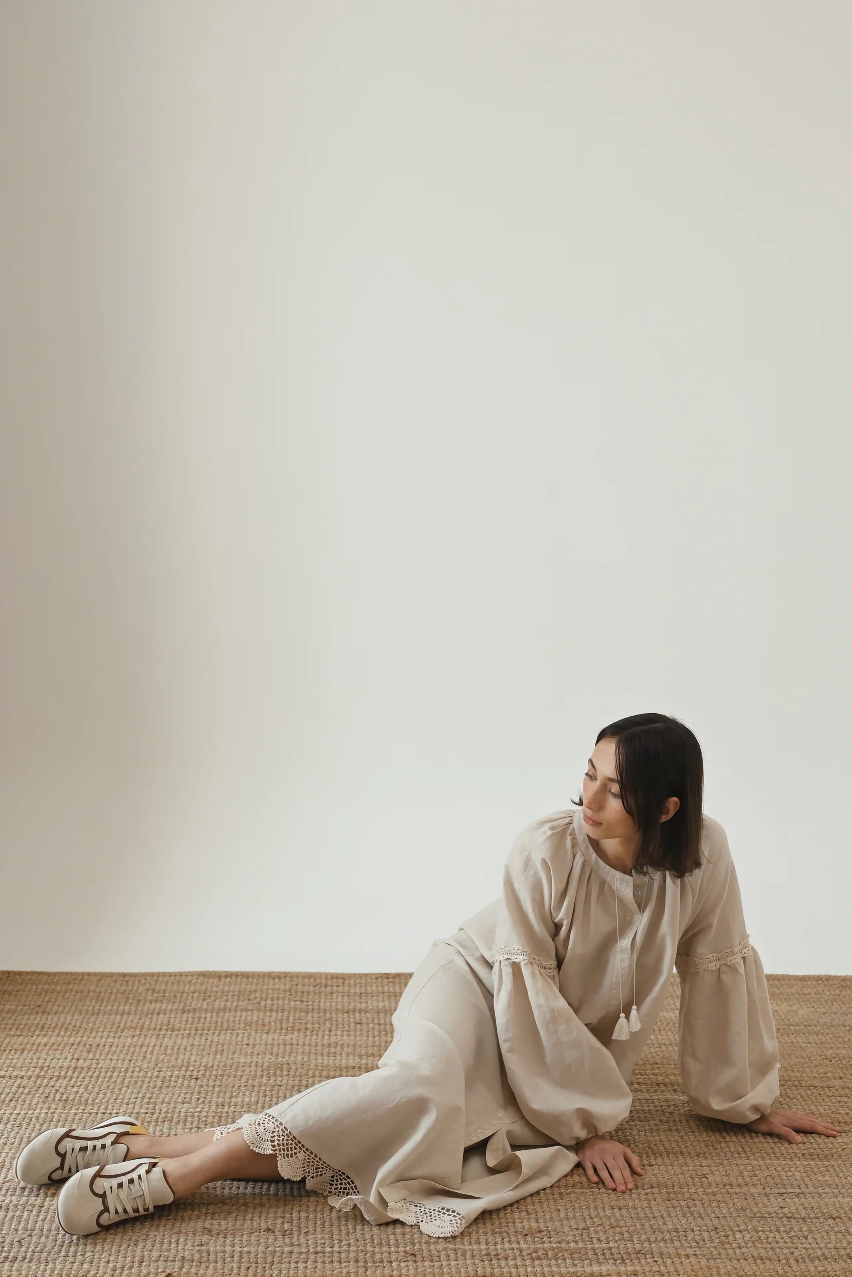 A woman wearing a beige outfit with voluminous sleeves and lace detailing, sitting on a woven rug and leaning back with one arm, looking to the side against a neutral background
