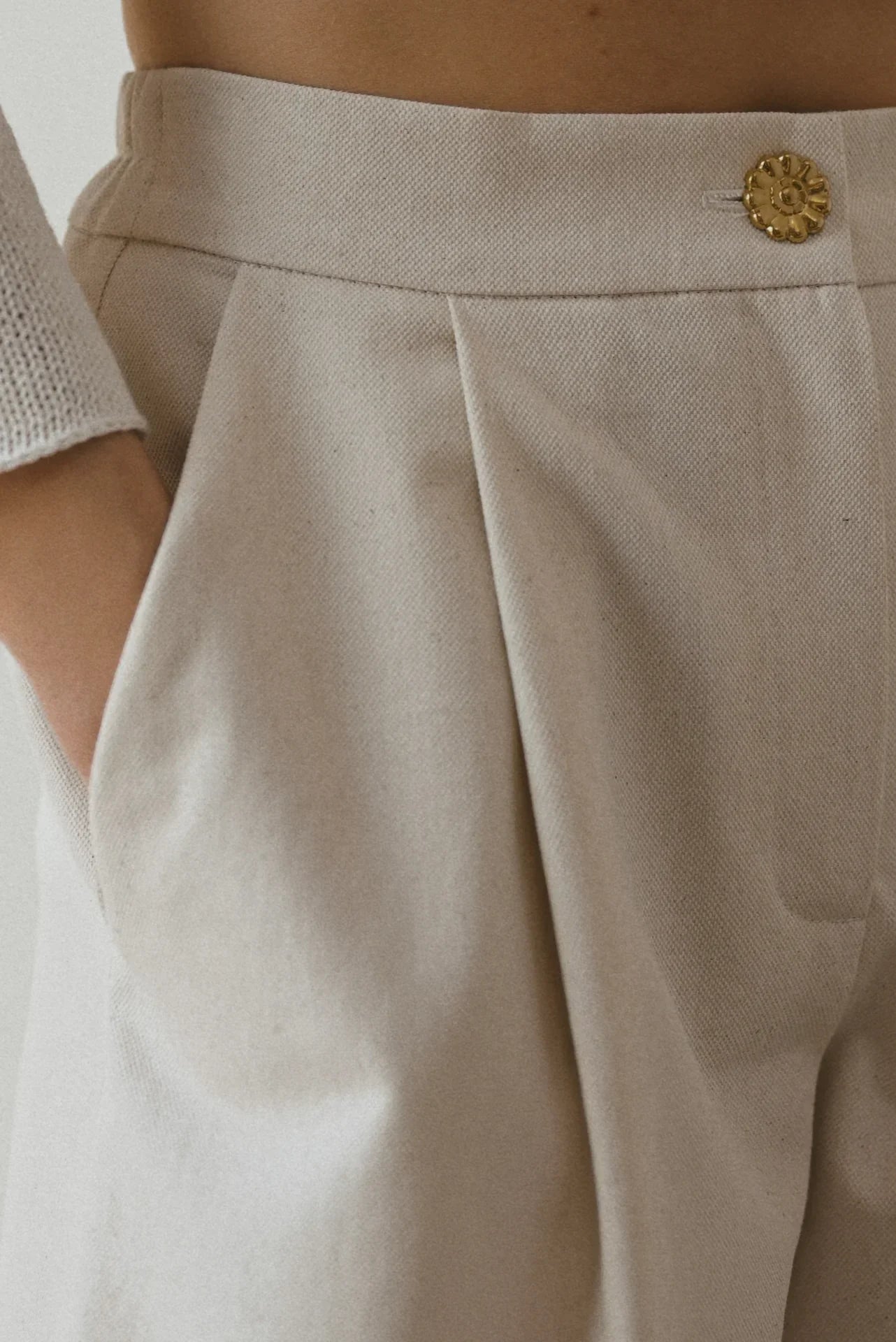 Close-up of a person wearing high-waisted beige trousers with pleats and a decorative button, with one hand in the pocket.