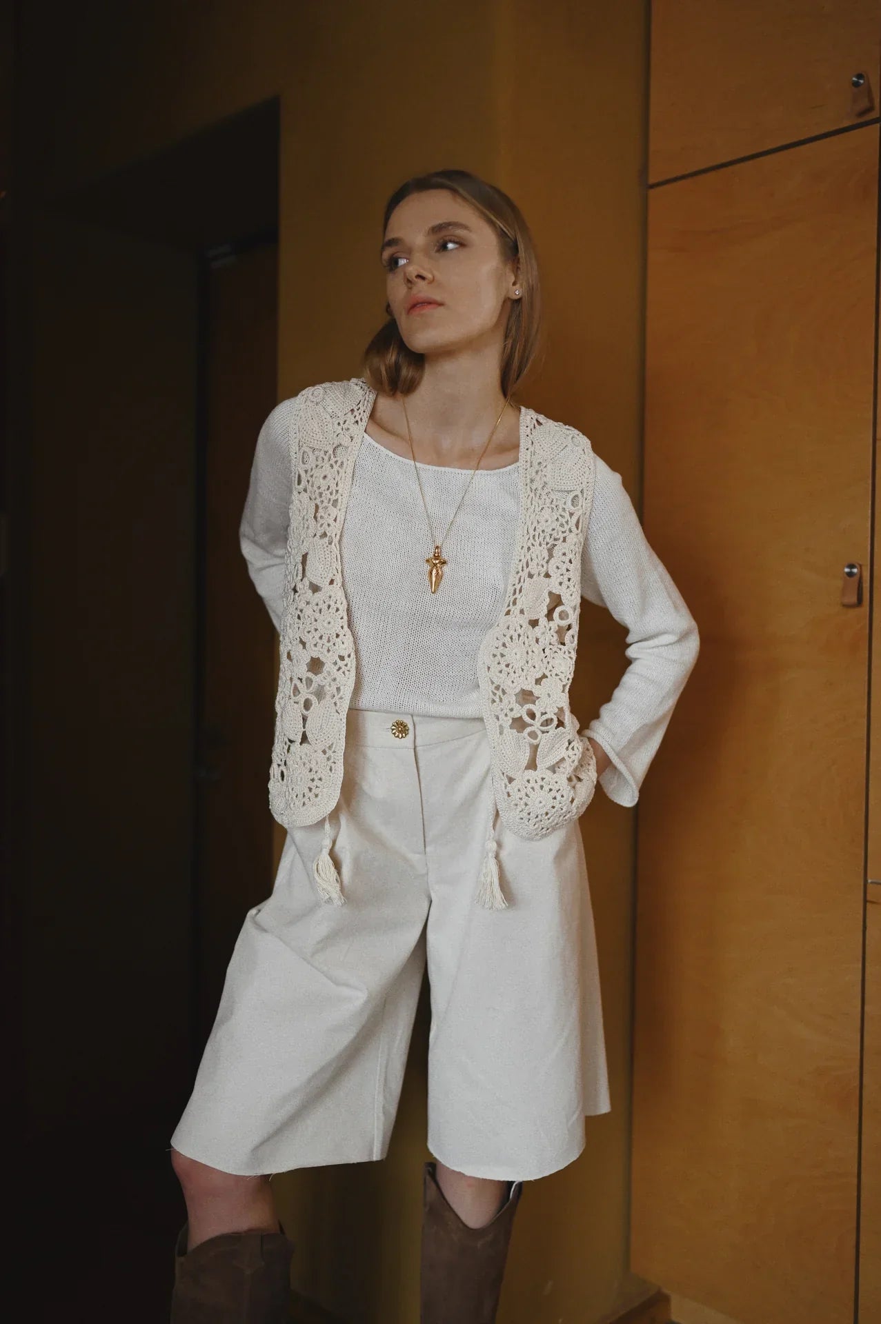 A woman wearing an off-white outfit, including a crocheted vest, high-waisted shorts, and a long necklace with a pendant, standing indoors against a warm-toned background.