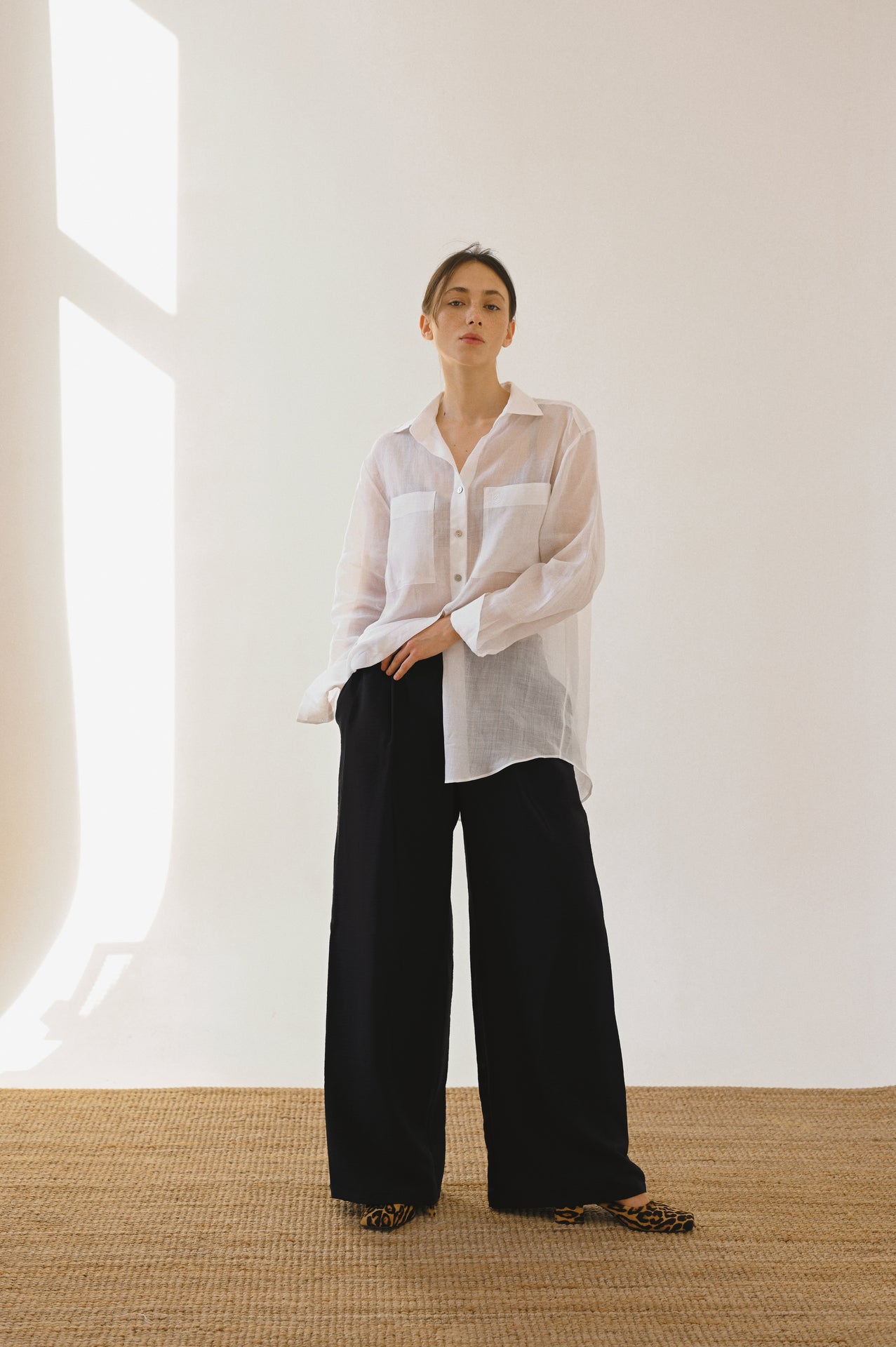 A woman wearing an oversized white button-up shirt and wide-leg black trousers, standing on a woven rug against a neutral background with sunlight streaming in