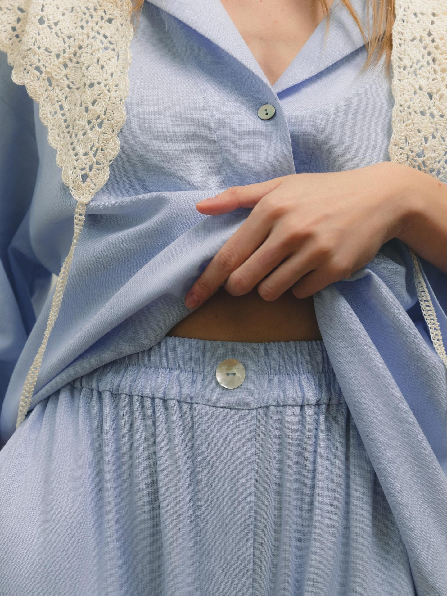 A close-up of a person wearing a light blue button-up shirt with a crocheted lace vest and matching high-waisted pants, gently lifting the hem of the shirt to reveal part of their waist.
