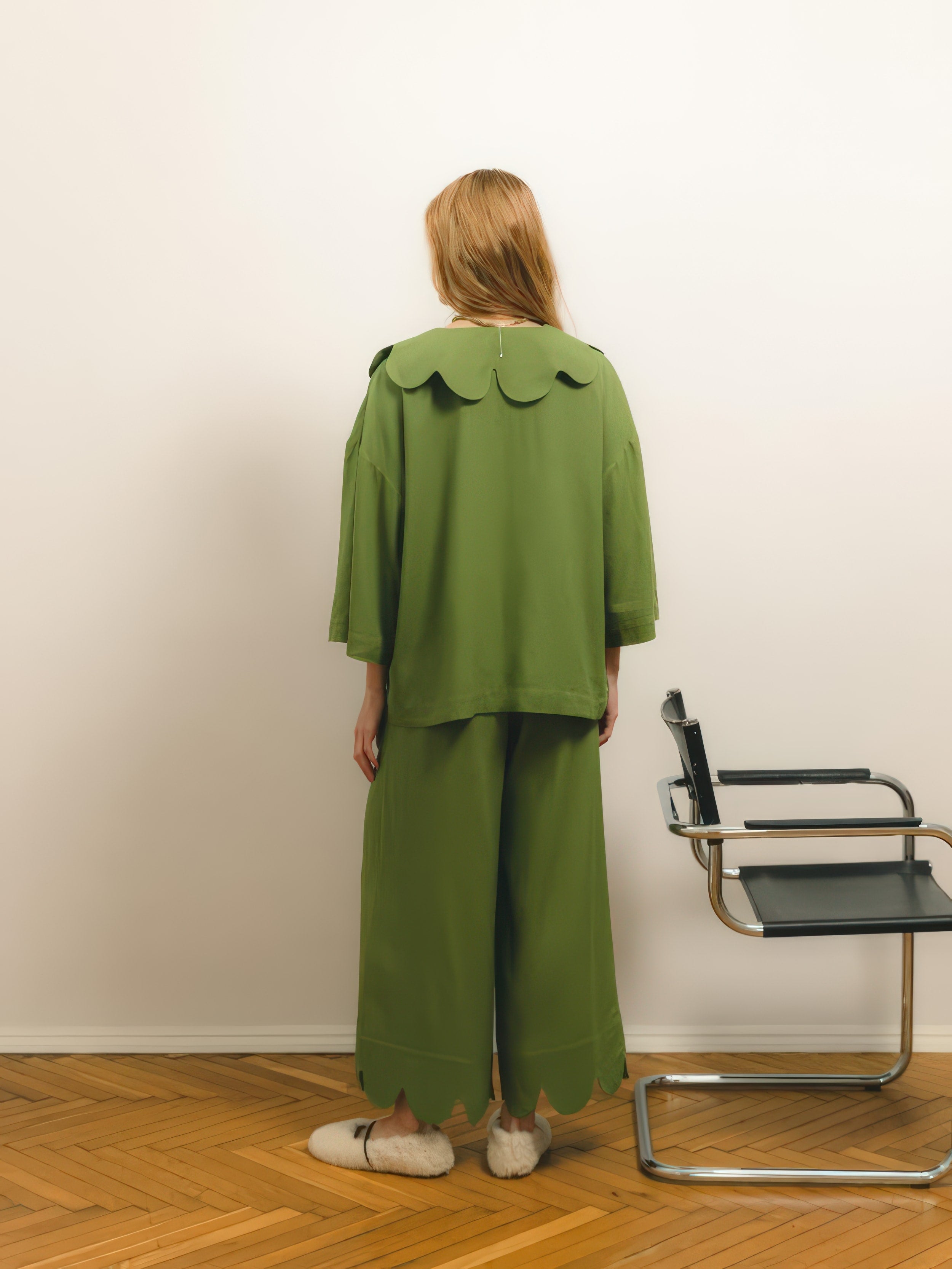 A woman with light brown hair standing with her back to the camera, wearing a loose green outfit with scalloped edges, next to a modern chair, against a neutral background.