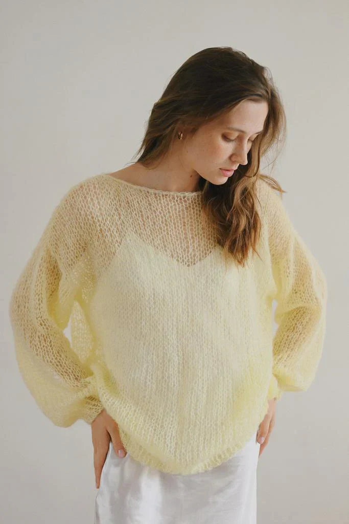 A woman with brown hair wearing a sheer yellow knit sweater over a white top, looking down with her hands in her pockets, against a neutral background.