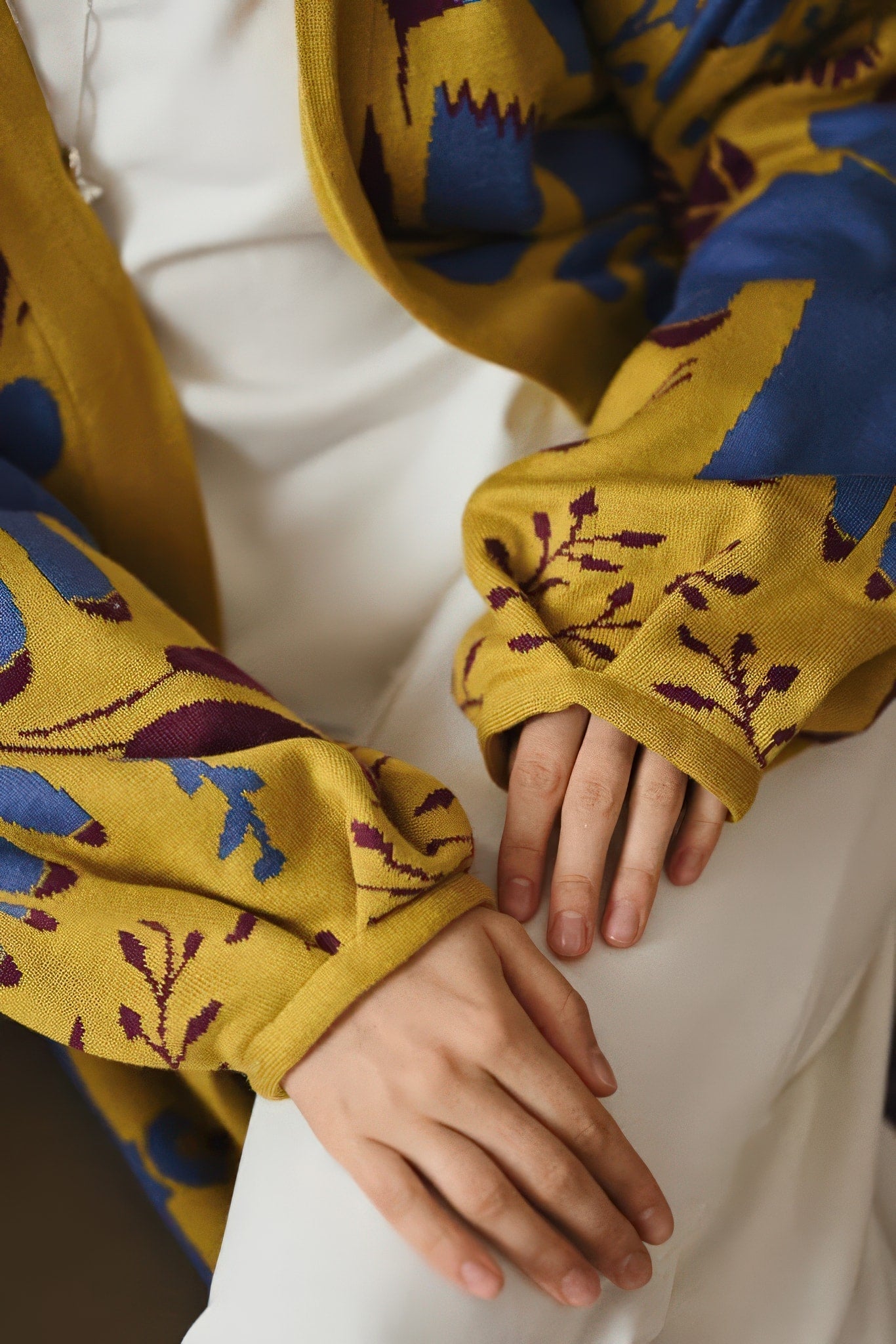 Close-up of a person’s hands resting on their lap, wearing a yellow garment with blue and burgundy floral patterns over a white outfit.