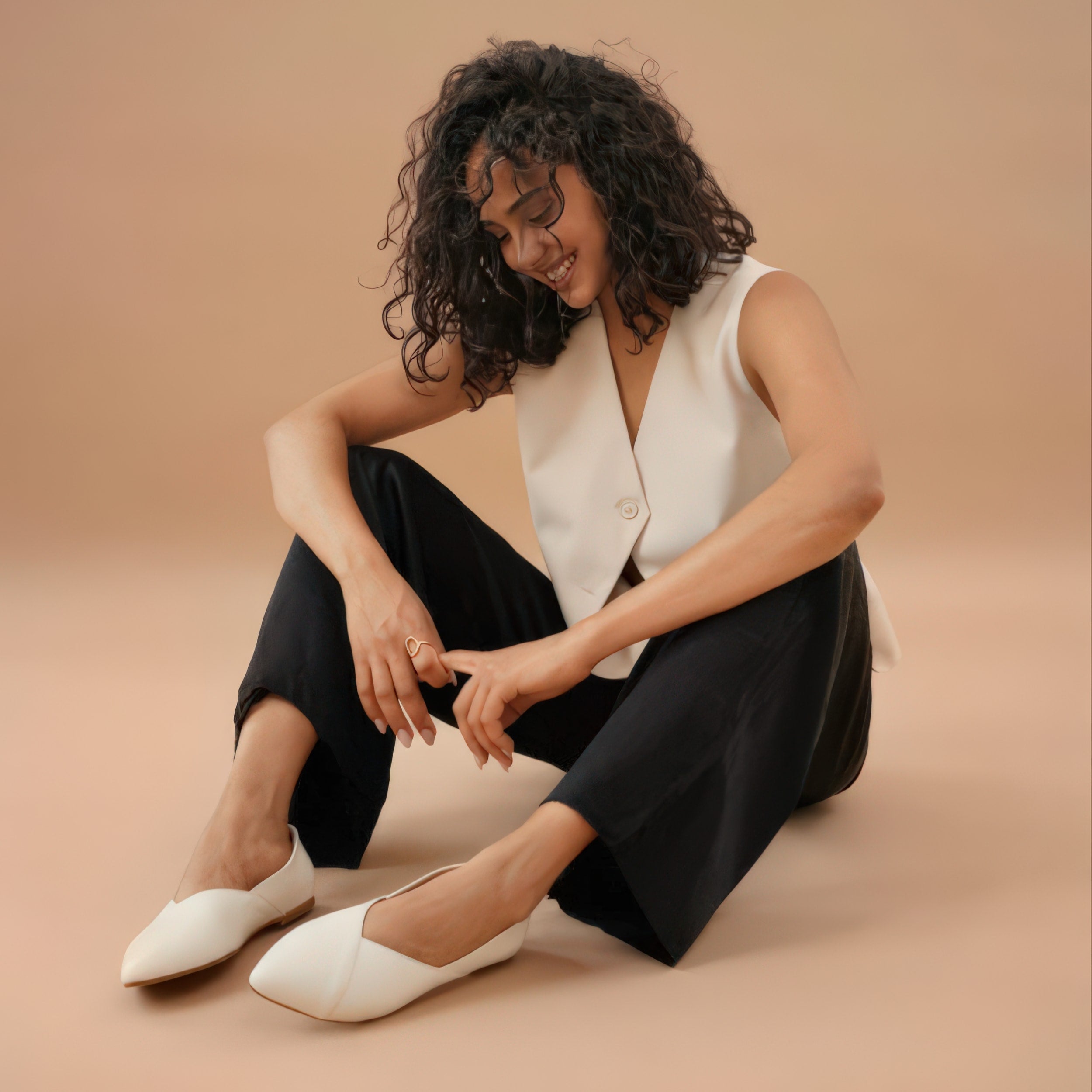 A woman with curly hair sitting on the floor against a beige background, wearing a sleeveless beige top, black pants, and white pointed-toe flats, smiling and looking downward