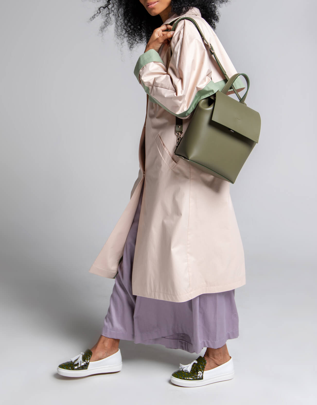 A woman wearing a light beige trench coat with green accents, a lavender skirt, white loafers with green details, and carrying an olive green backpack, walking against a gray background.