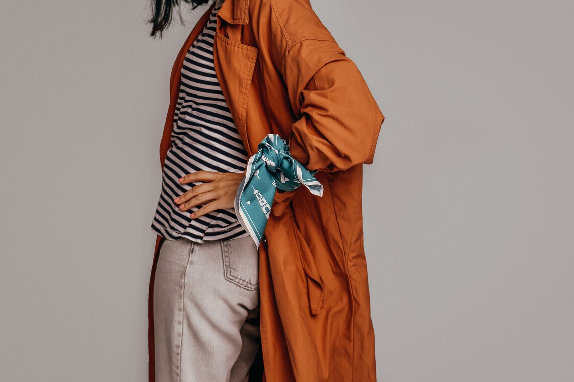 A person wearing a rust-colored trench coat over a striped top and beige pants, with a teal patterned scarf tied around their wrist, posing against a neutral background