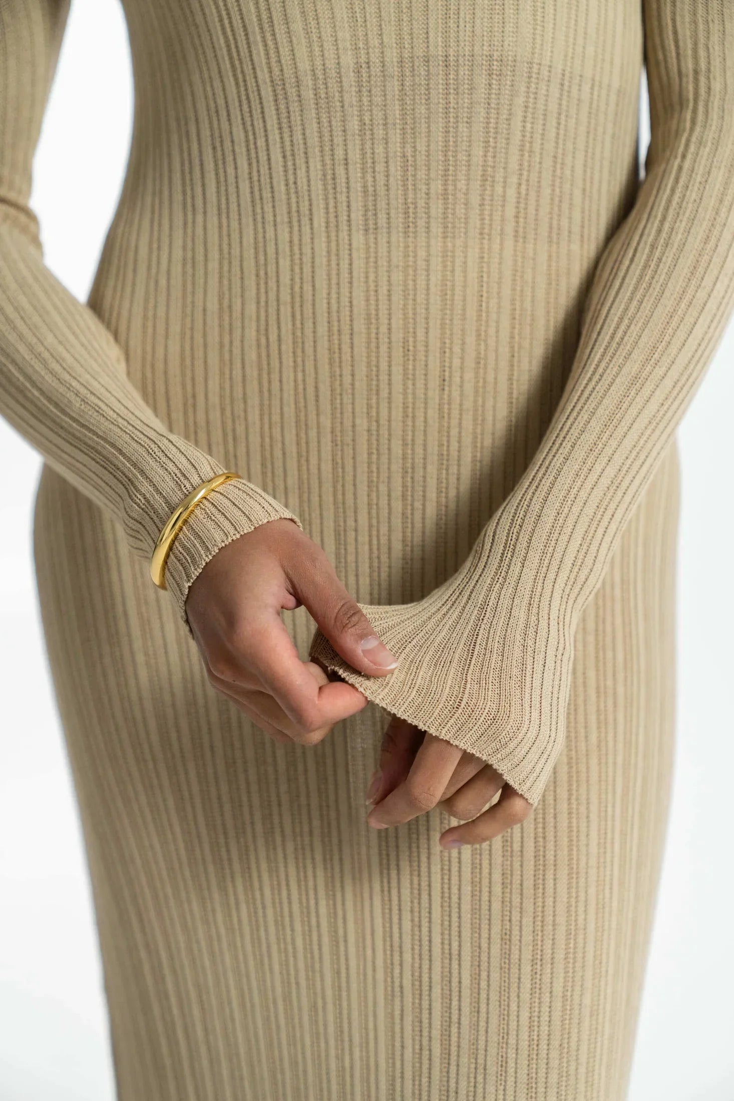 A close-up of a person wearing a beige ribbed knit dress with long sleeves, showcasing their hands adorned with a gold bracelet and a ring.