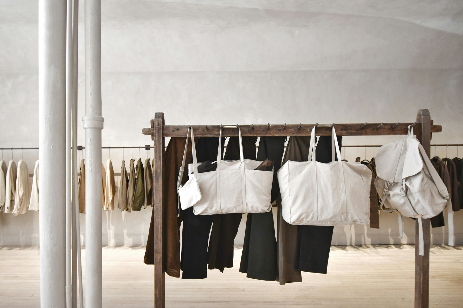 A minimalist store interior featuring a wooden clothing rack displaying neutral-toned garments and large beige canvas bags, with additional clothing displayed on racks in the background.
