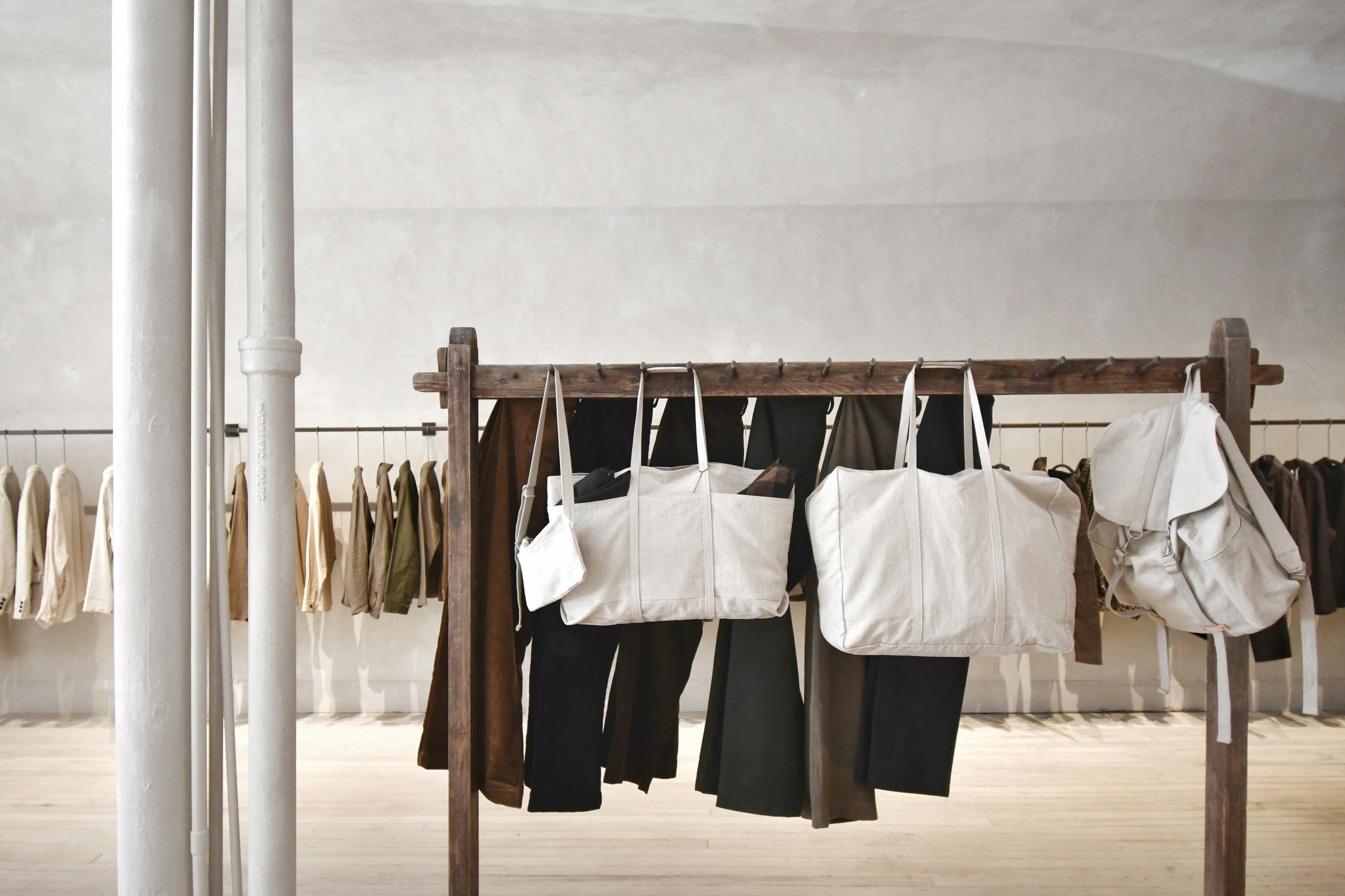 A minimalist store interior featuring a wooden clothing rack displaying neutral-toned garments and large beige canvas bags, with additional clothing displayed on racks in the background.