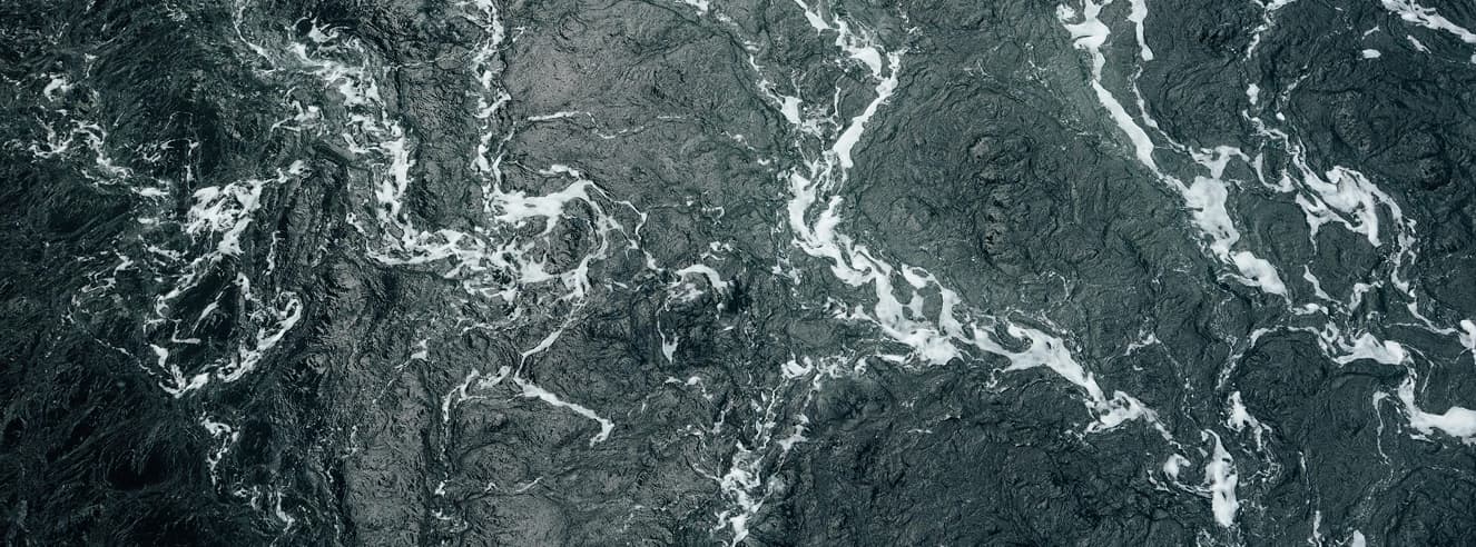 A close-up view of dark turbulent water with white foam patterns swirling across the surface, creating a dramatic and textured appearance.